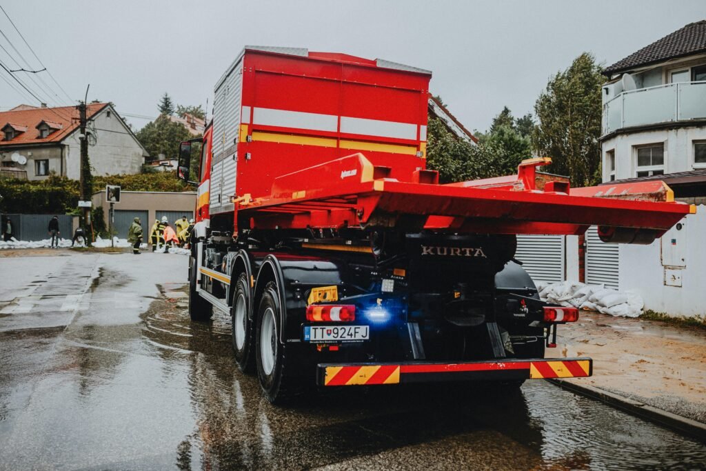 A red tow truck parked on a wet street in an urban neighborhood with workers in the background.